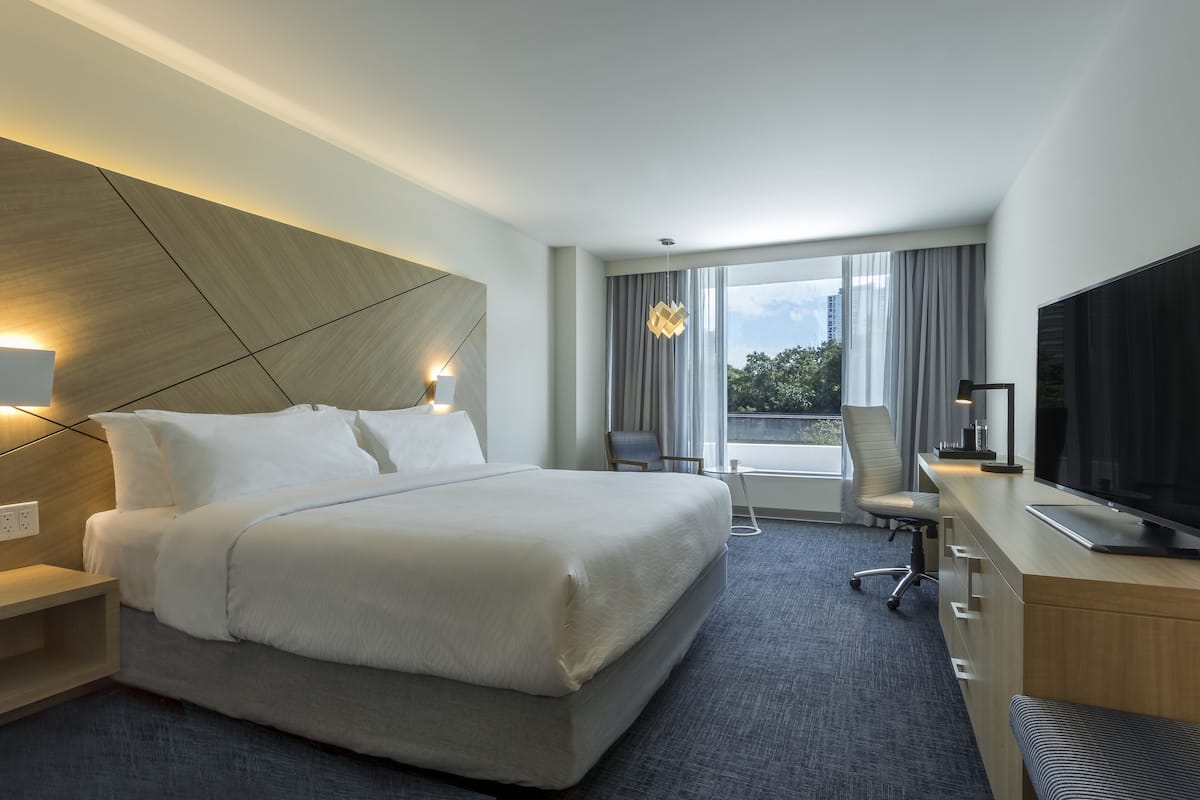 Habitación de hotel moderna con una cama grande, un escritorio y un televisor de pantalla plana, y vistas al exterior a través de amplias ventanas.