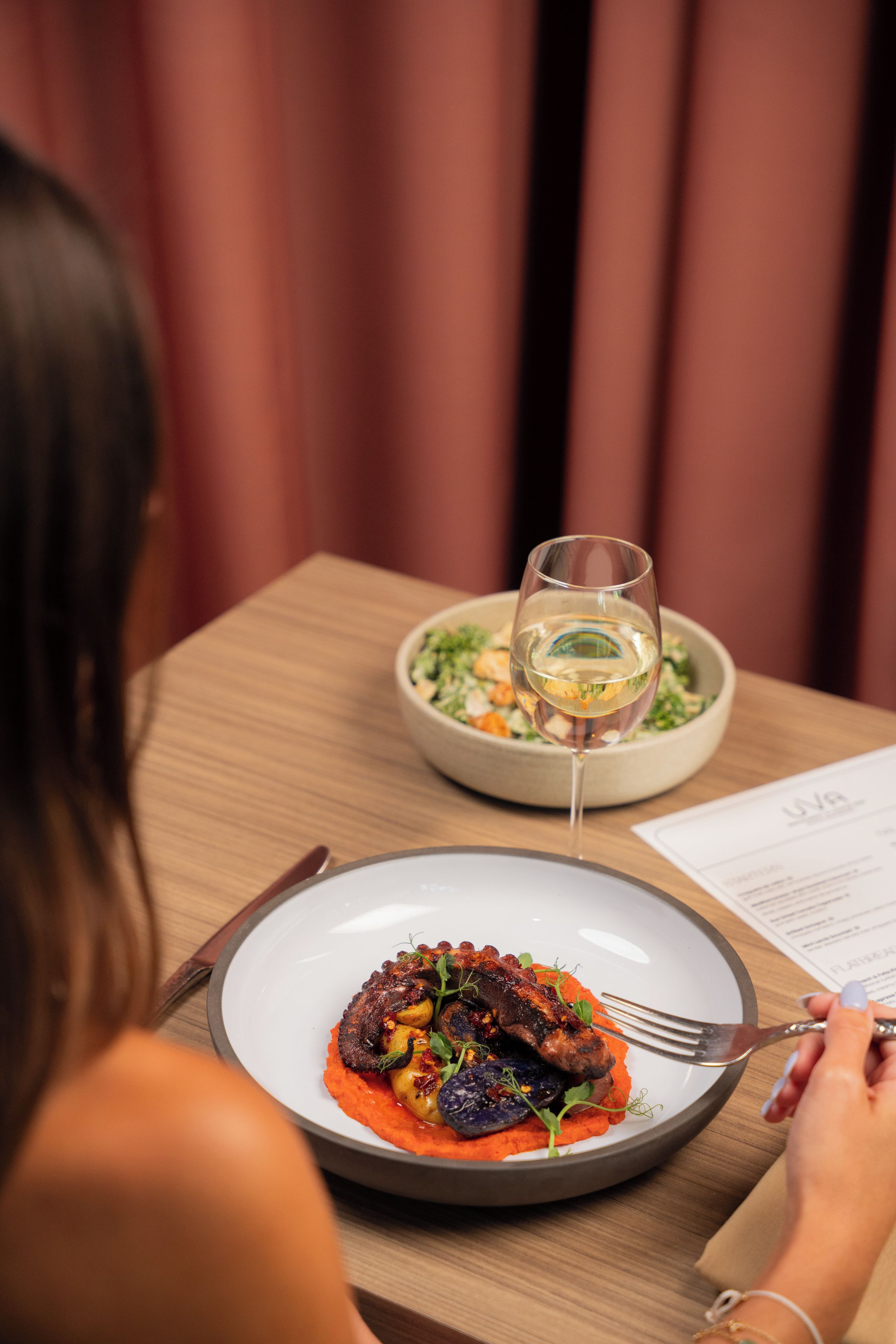 A dining table with a plate of food, a glass of white wine, and a menu.