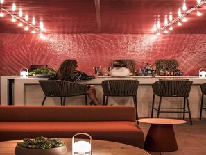 A bar area with a red textured wall, bar stools, a counter with a person behind it, and string lights.