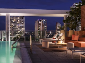 Zona de piscina en la azotea con sillones, un sofá y vista a edificios de la ciudad de noche.