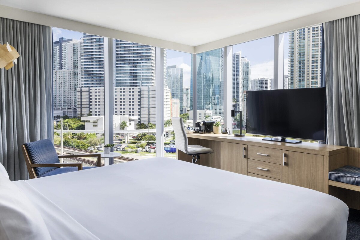 Habitación de hotel con una cama grande, un escritorio con televisor y vistas a edificios altos.