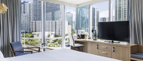 A hotel room with a large bed, a desk with a television, and a view of tall buildings.