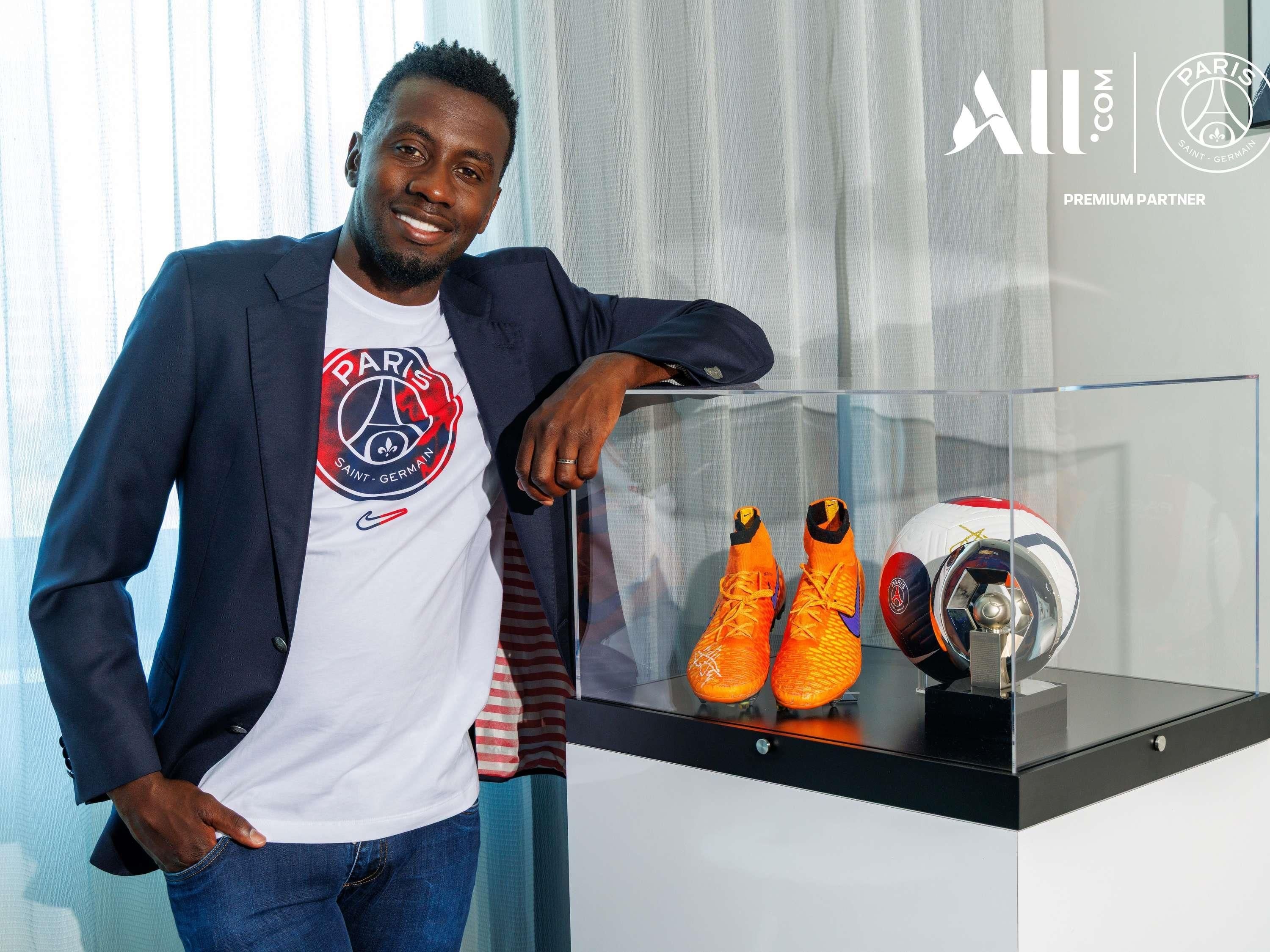 A man in a Paris Saint-Germain t-shirt standing next to a display case with a soccer ball and shoes.