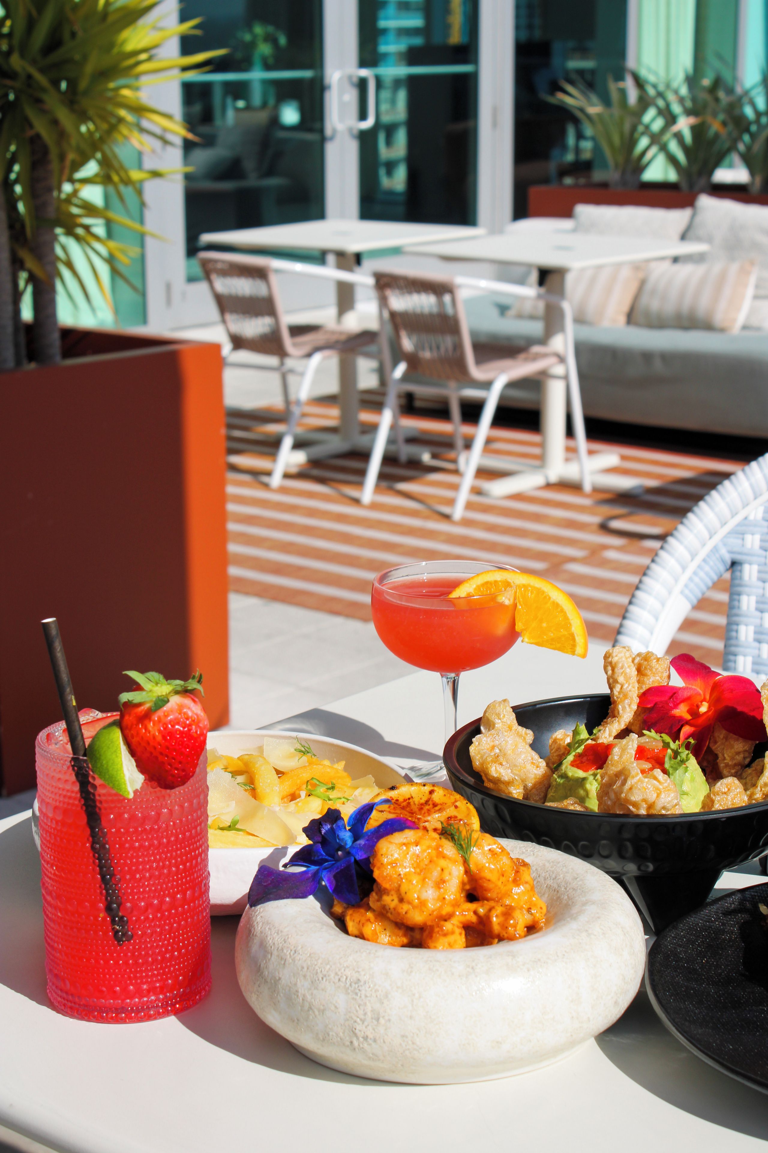 A dining table with a variety of food and drinks, including a red cocktail with a strawberry garnish, a bowl of shrimp, and a plate with a white dessert.