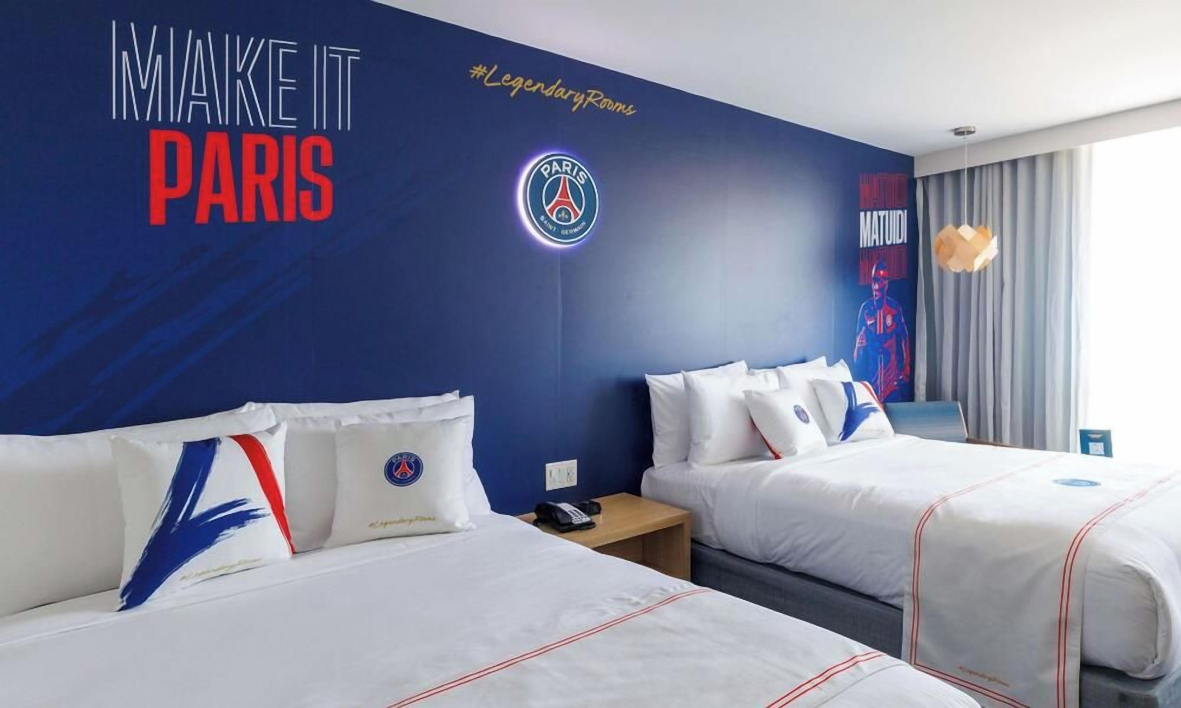 A hotel room with two beds, a wall with a Paris 2024 logo, and a window with curtains.