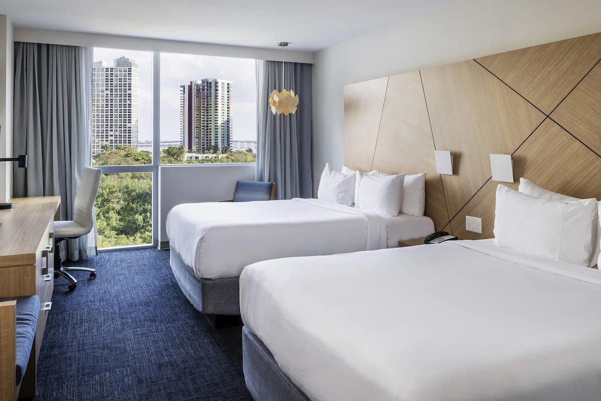 Habitación de hotel con dos camas, un escritorio, una silla y vistas a edificios altos.