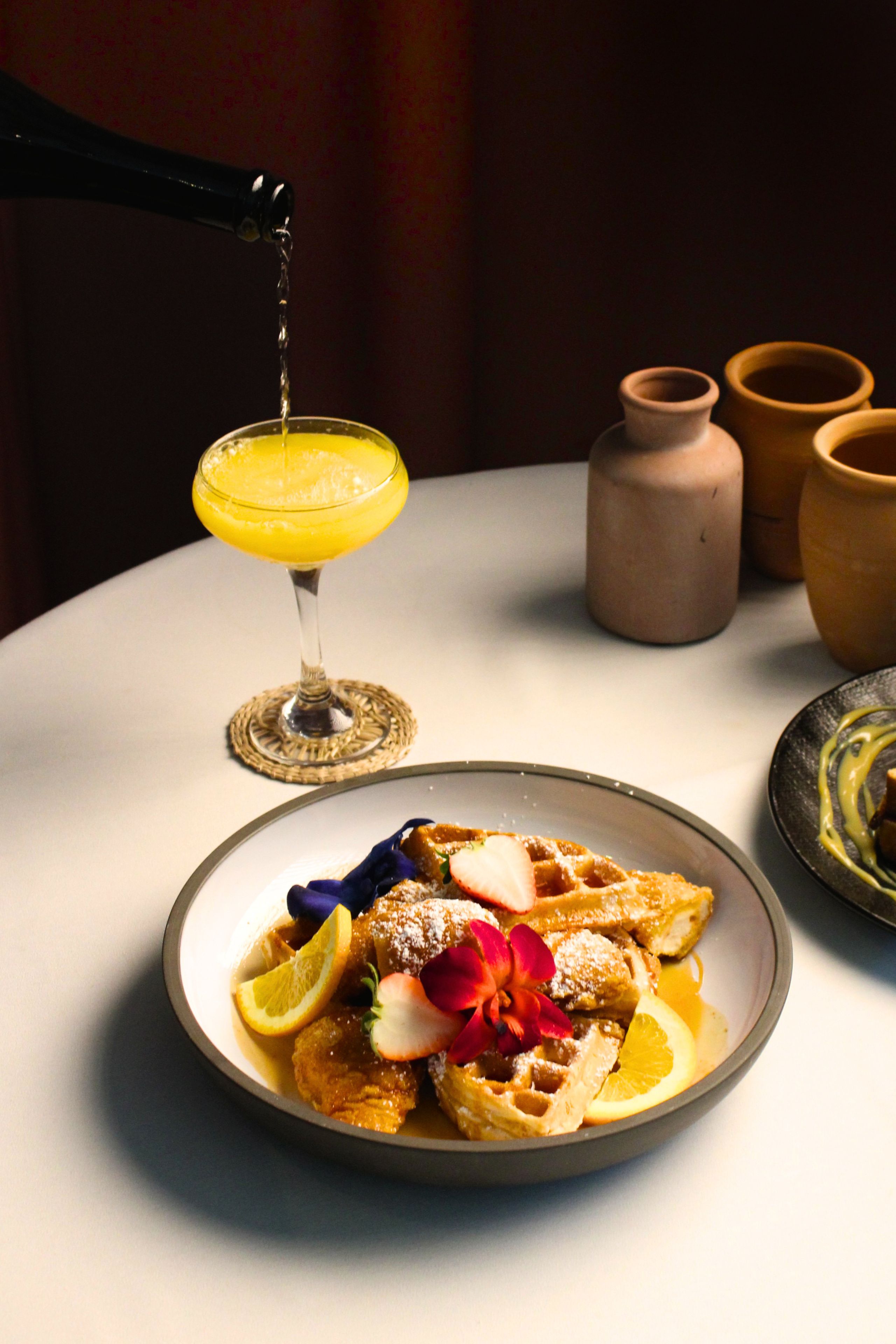 A table with a plate of food, a glass of a yellow beverage, and a vase with flowers.