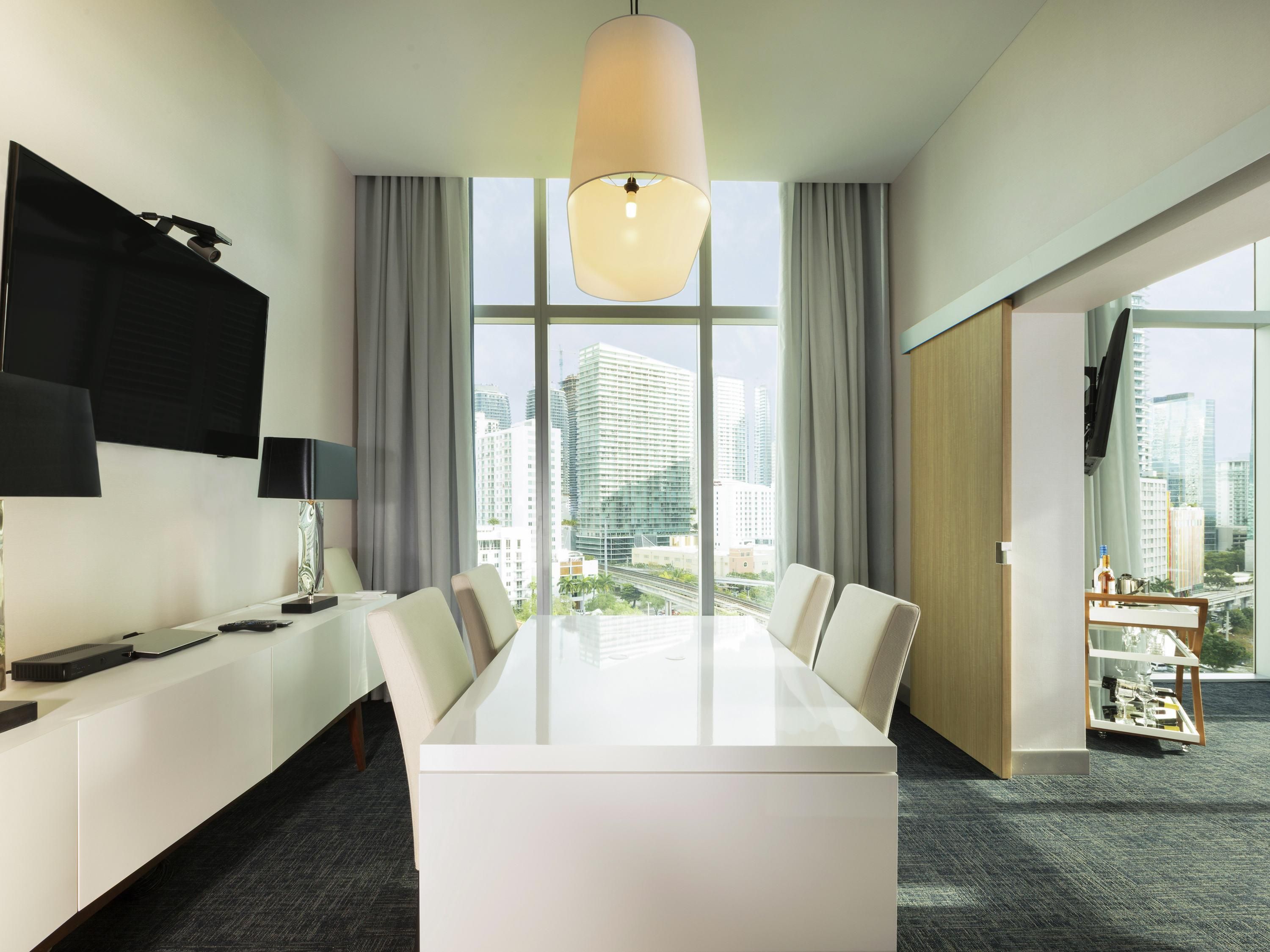 Een moderne hotelkamer met een grote eettafel, een flatscreen televisie, een lamp en uitzicht op de stad.