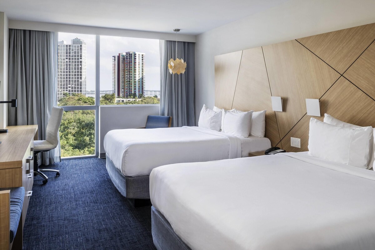 Habitación de hotel con dos camas, un escritorio, una silla y vistas a edificios altos.