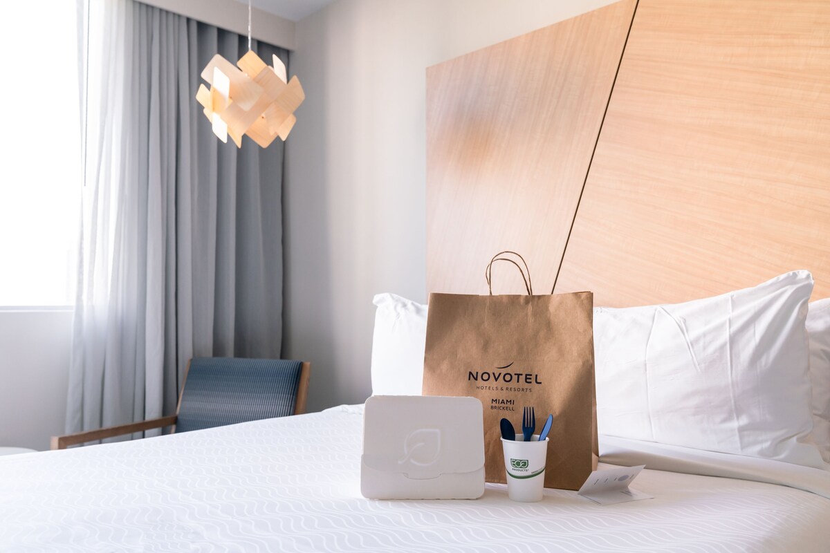 Habitación de hotel con cama, laptop, vaso de papel con cubiertos y bolsa marrón con el logotipo de Novotel.