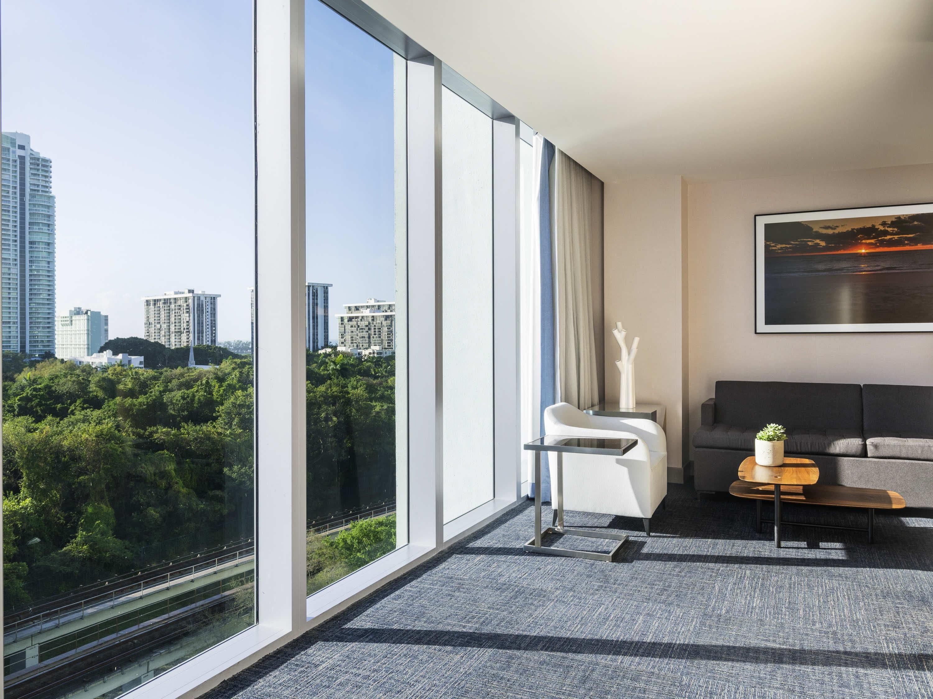 A modern hotel room with large windows, a sofa, a small table, and a painting.