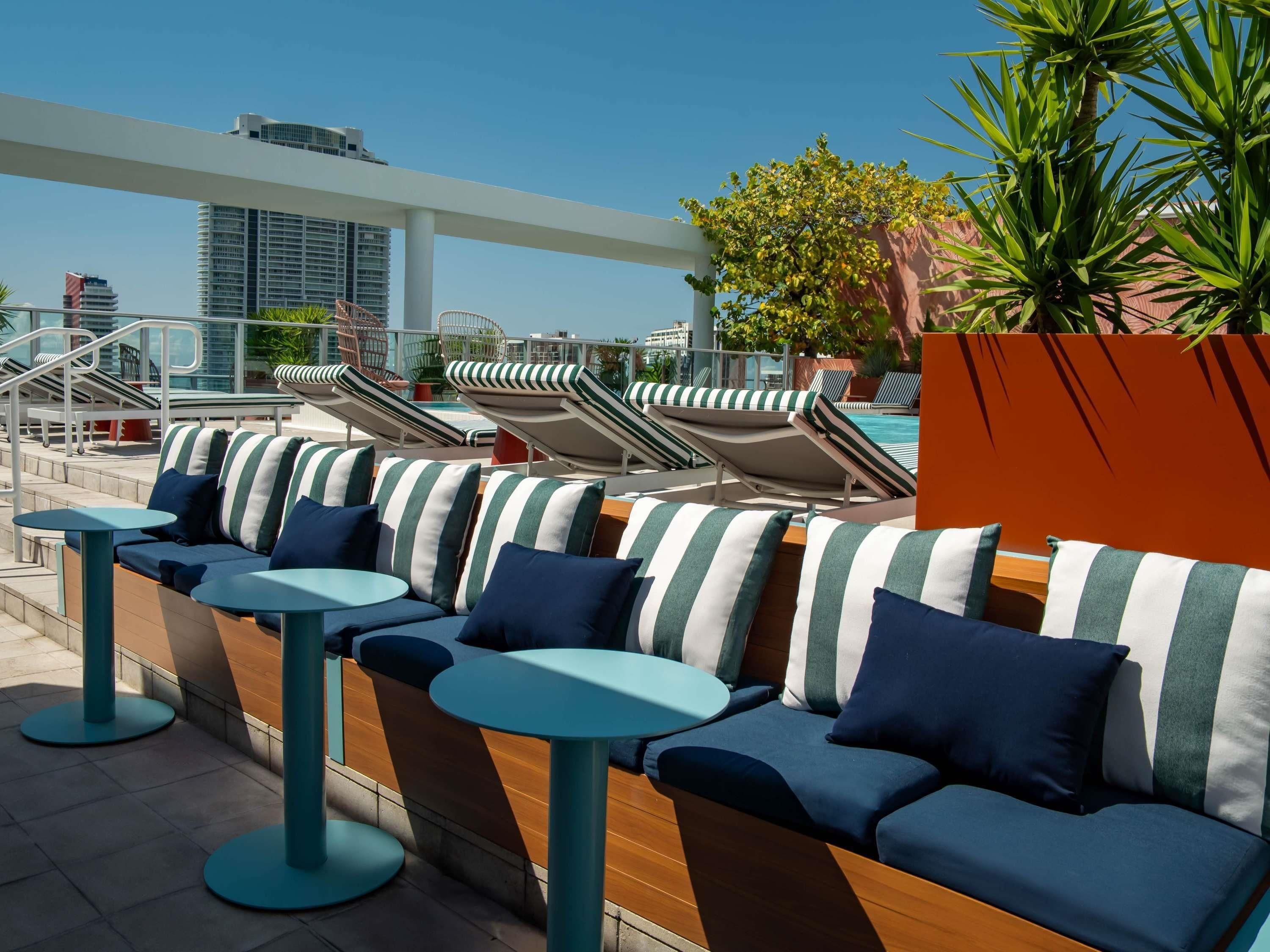 A rooftop lounge with lounge chairs and tables, a pool, and a view of the city skyline.