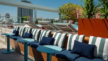 A rooftop lounge with lounge chairs and tables, a pool, and a view of the city skyline.