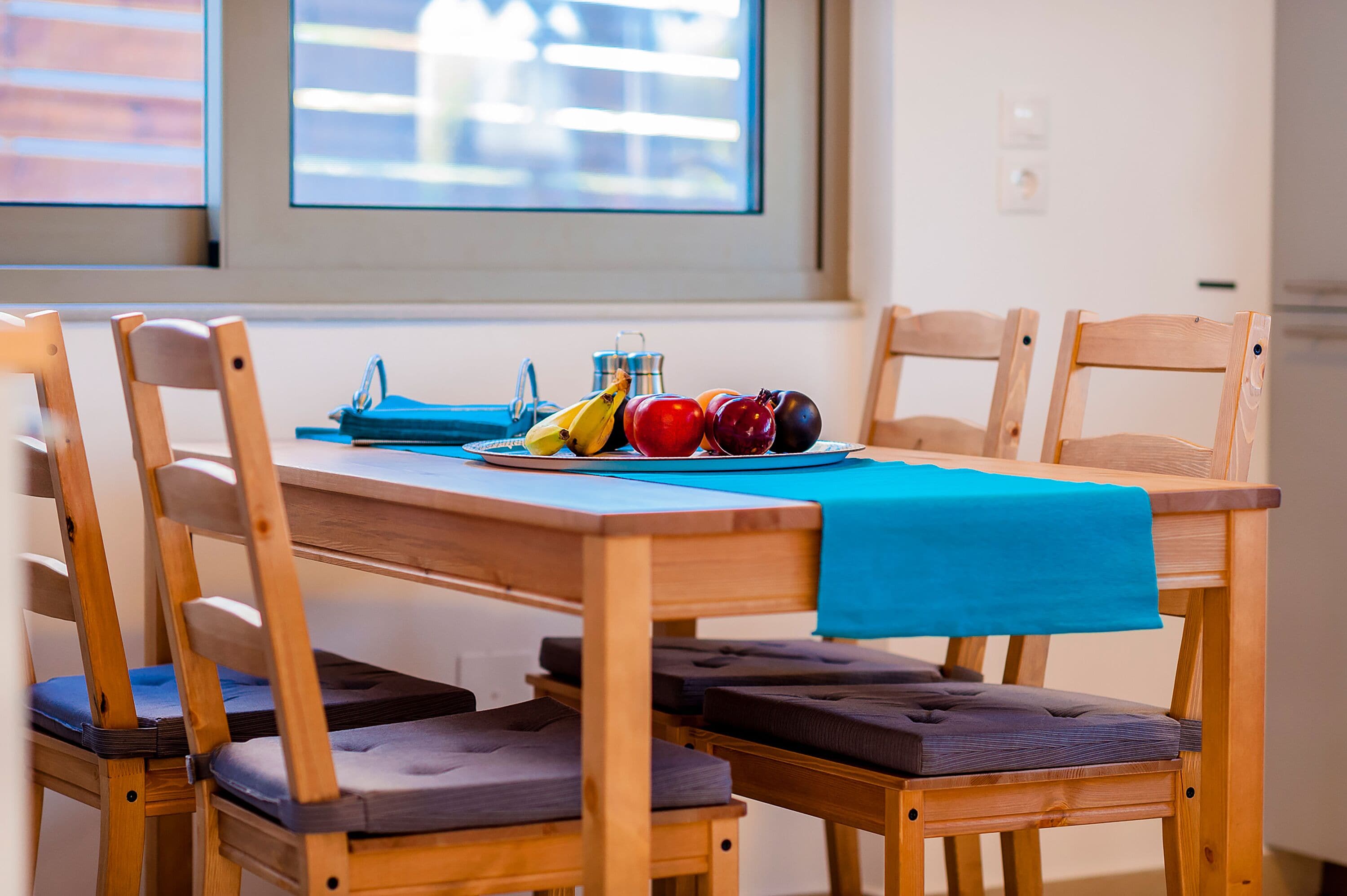 Une table de salle à manger en bois, recouverte d’une nappe bleue, entourée de chaises en bois dotées d’assises rembourrées.