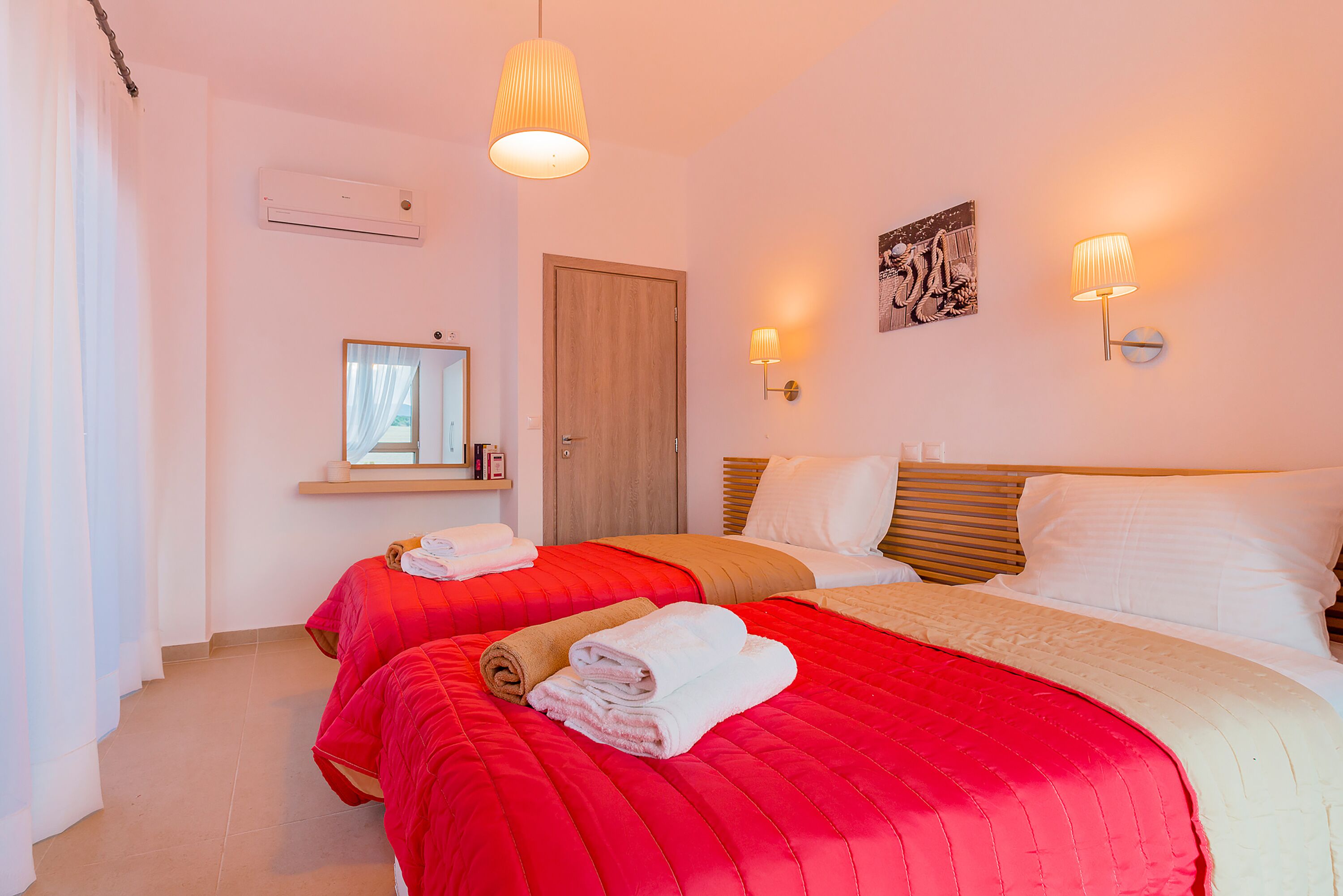 A hotel room with two beds, each with red and beige bedding, a wooden door, a mirror, and wall-mounted lights.