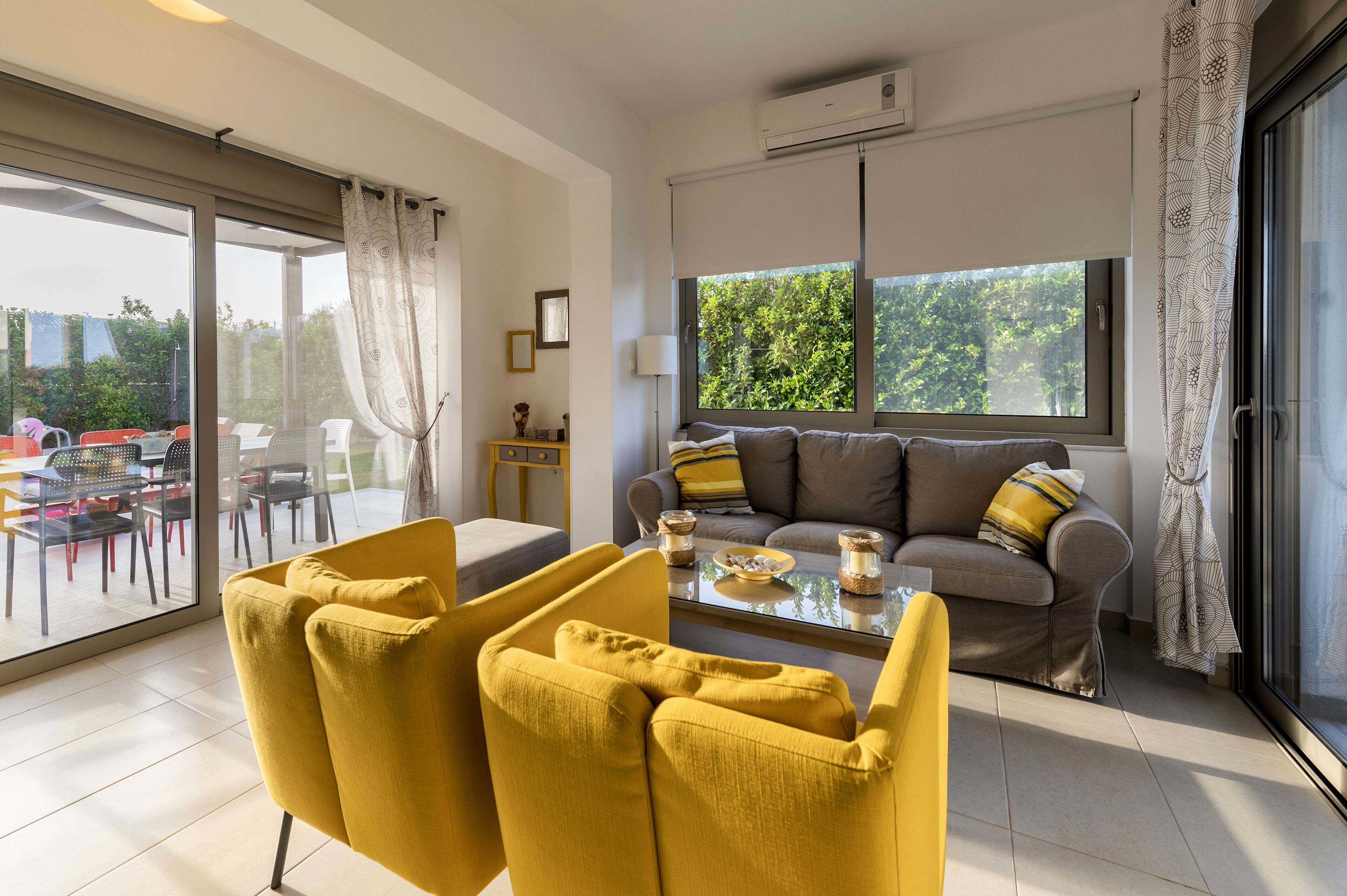A modern living room with a sofa, yellow armchairs, a glass coffee table, and a large window with curtains.