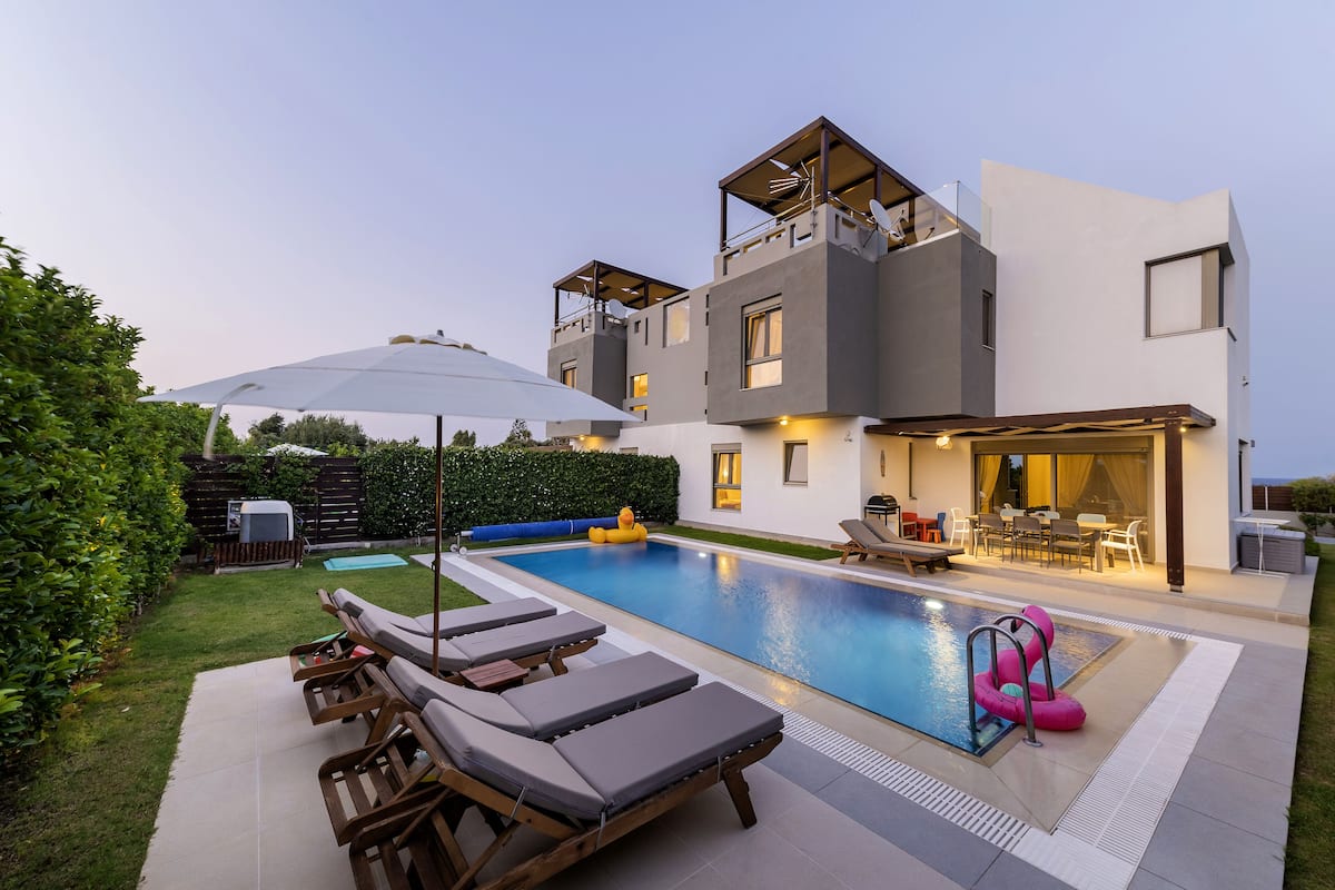 Een modern huis met twee verdiepingen, een zwembad, terrasmeubels buiten en een overdekte patio.