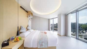 A modern hotel room with a large bed, a bedside table with fruits, a nightstand, and a large window offering a city view.