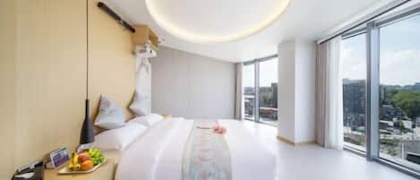 Una habitación de hotel moderna con una cama grande, una mesita de noche con frutas, una mesita auxiliar y un amplio ventanal con vistas a la ciudad.