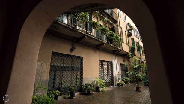 An arched passageway with a building on one side, featuring balconies with potted plants and a cobblestone ground. - Italianway   - Argelati (Milan)
