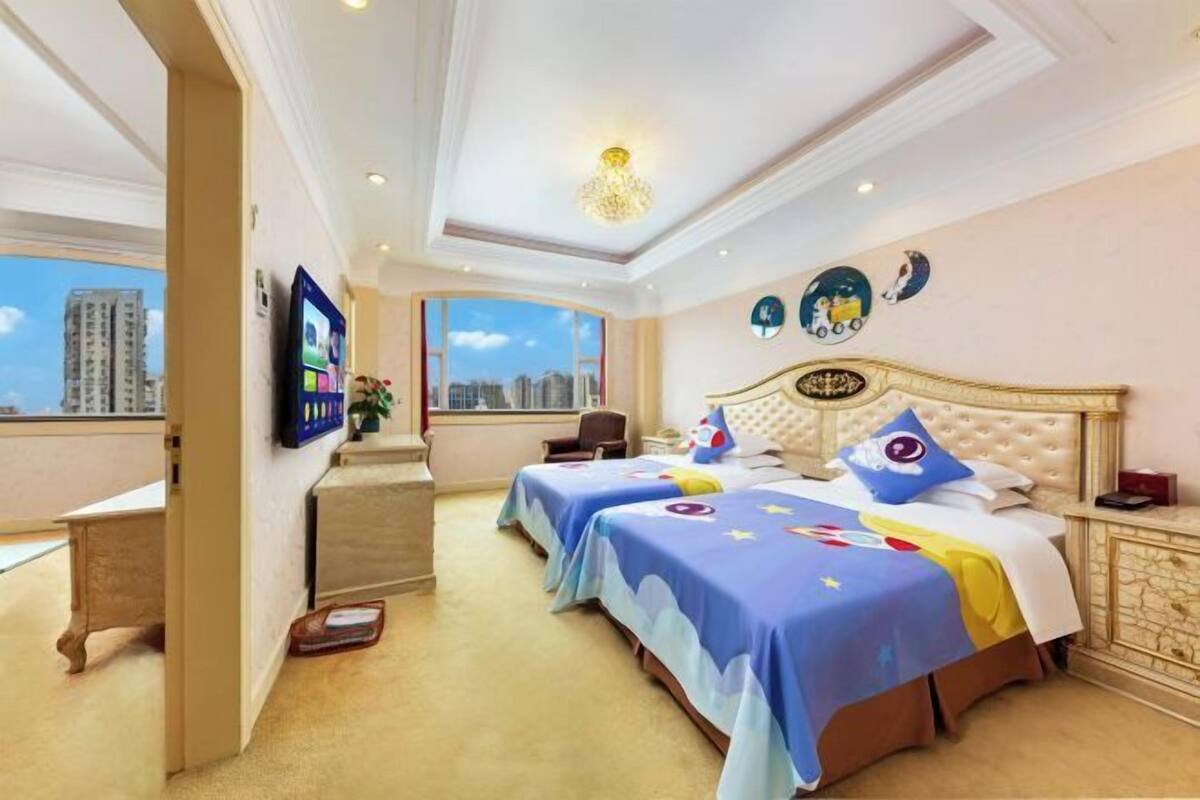 Habitación de hotel con dos camas, un ventanal con vistas a la ciudad, una araña y un televisor en la pared.