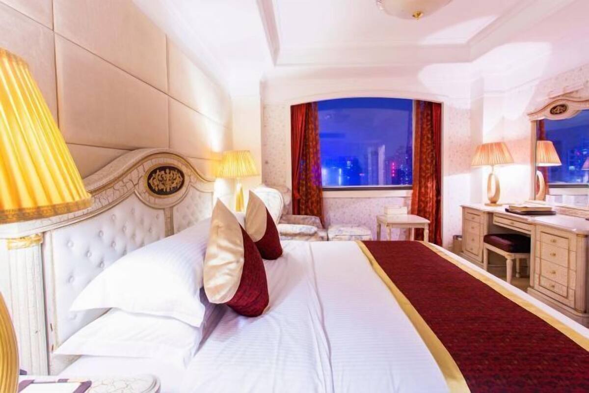 Habitación de hotel con una cama grande, dos mesitas de noche con lámparas, un escritorio y vistas a la ciudad de noche.