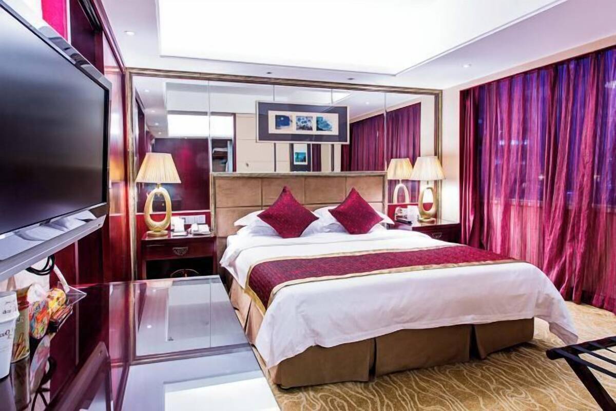 Habitación de hotel con una cama grande, dos mesitas de noche con lámparas, un televisor de pantalla plana, un espejo y un ventanal con cortinas.