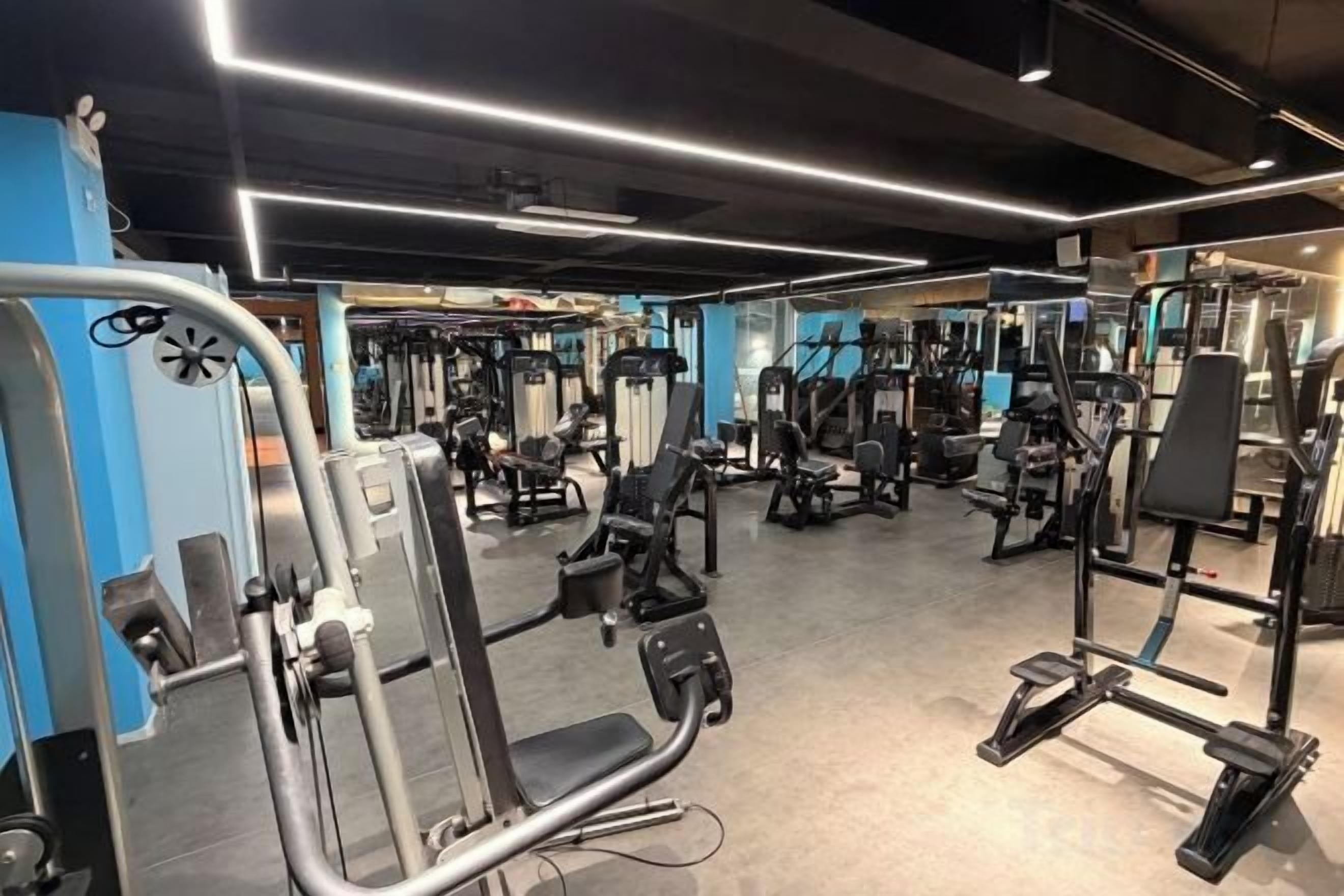Un gimnasio con diversas máquinas de ejercicio, espejos e iluminación lineal en el techo.