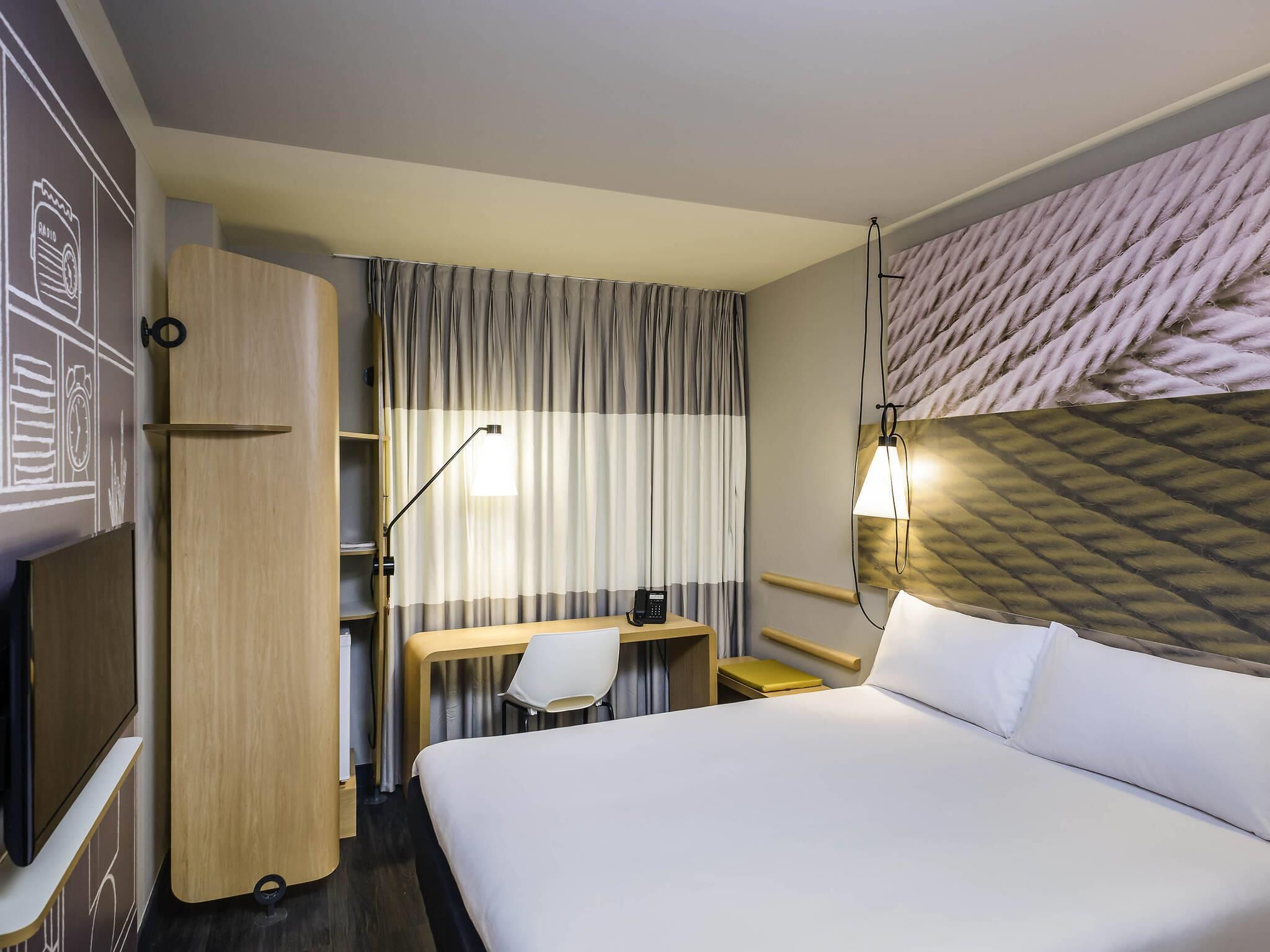 A hotel room with a bed, a desk, a chair, a wardrobe, and a wall-mounted lamp.
