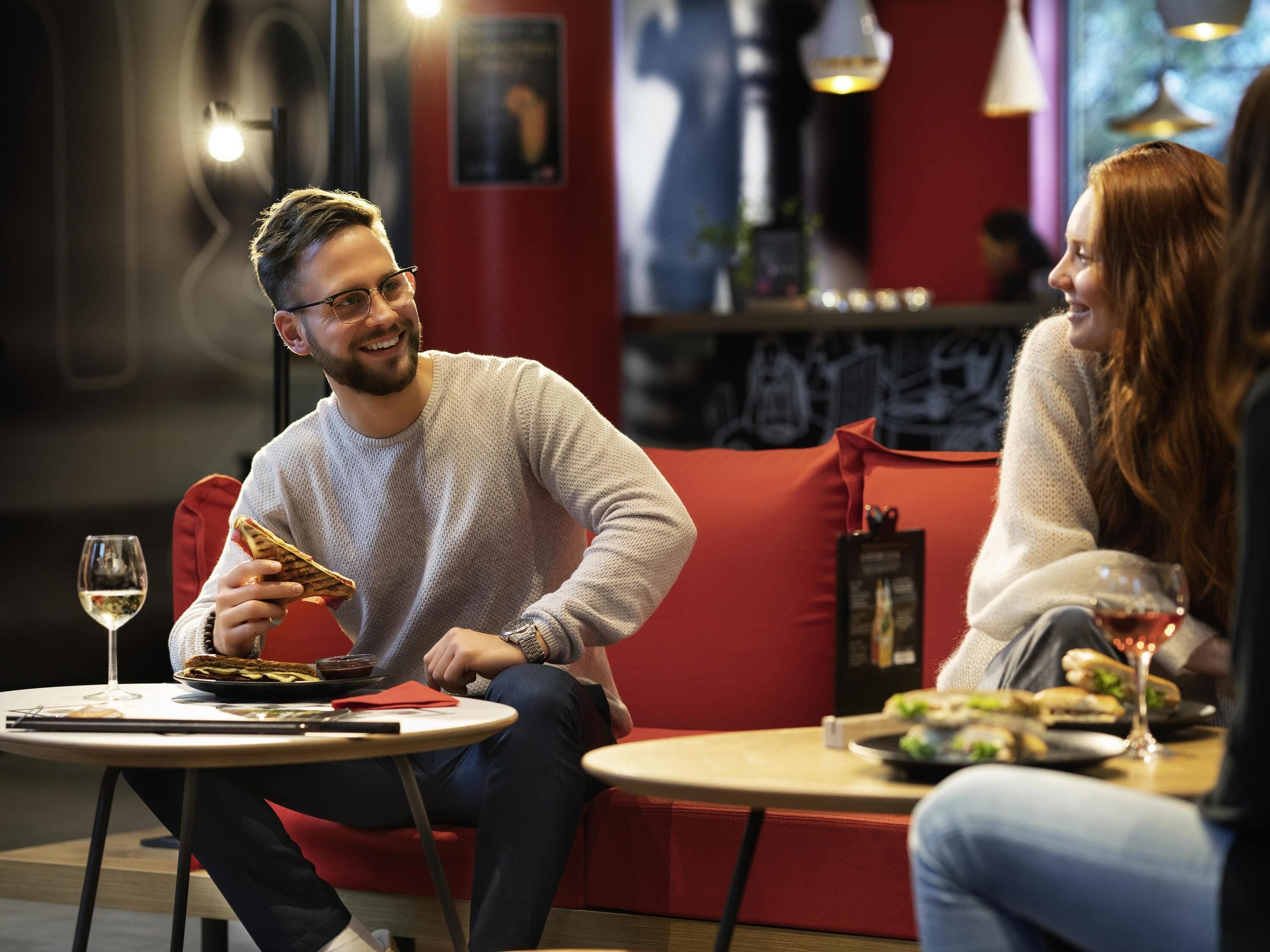 Une personne est assise sur un canapé rouge. Devant elle, sur une petite table ronde, se trouvent une assiette de nourriture et un verre de vin.