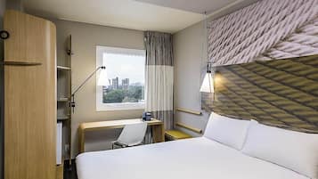 Quarto de hotel com cama, escrivaninha, cadeira e vista para a cidade pela janela.