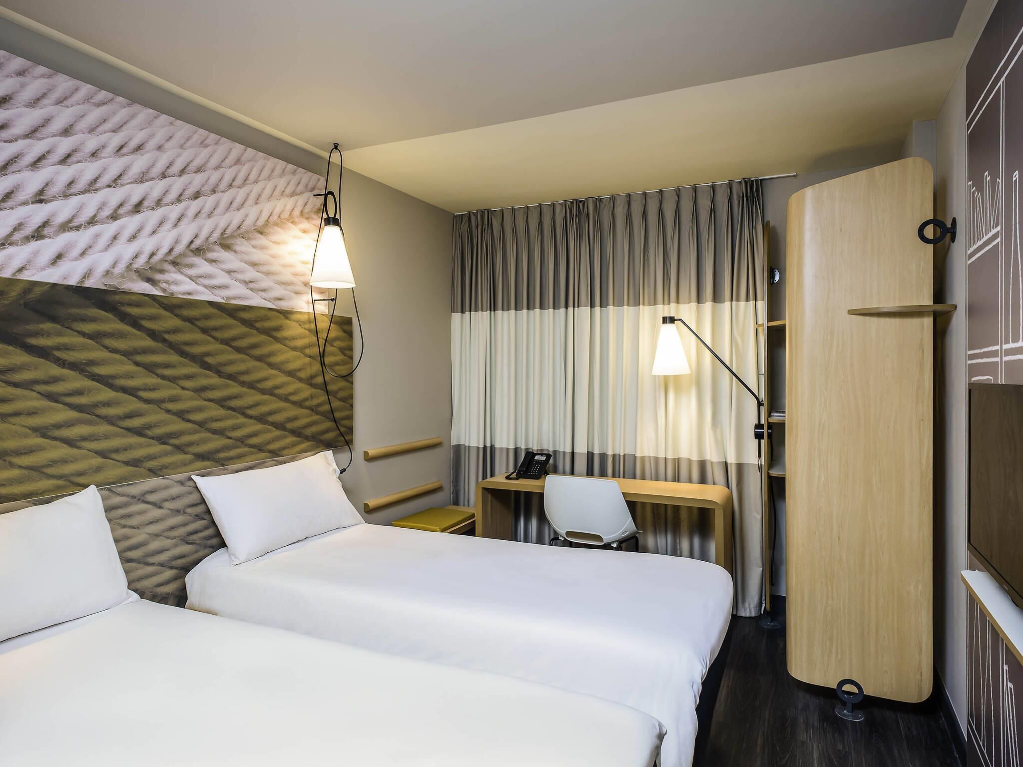 A hotel room with two beds, a desk, a chair, a wardrobe, and a headboard with a textured design.