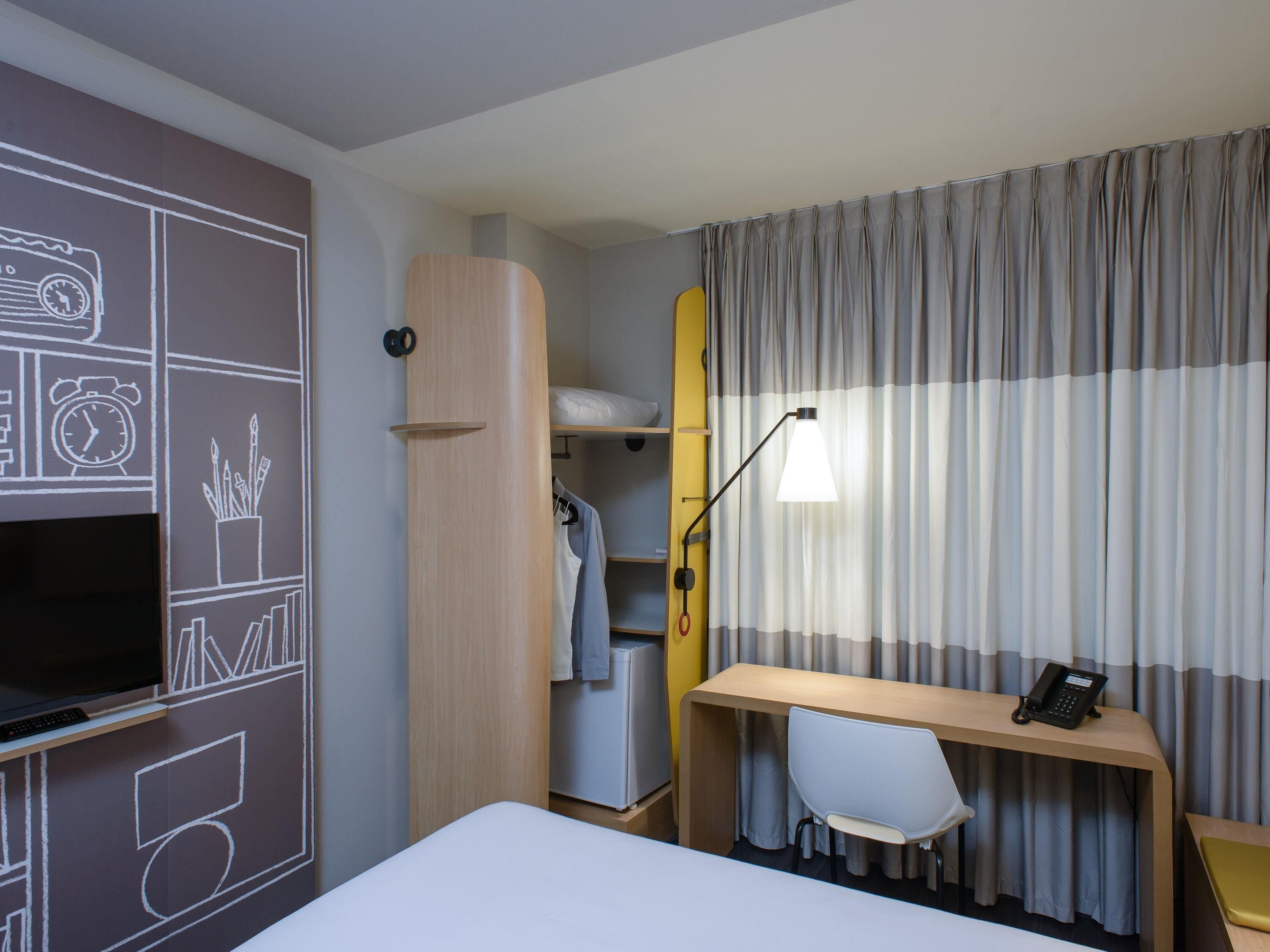 A hotel room with a bed, a desk with a chair, a TV, a wardrobe, and a window with curtains.