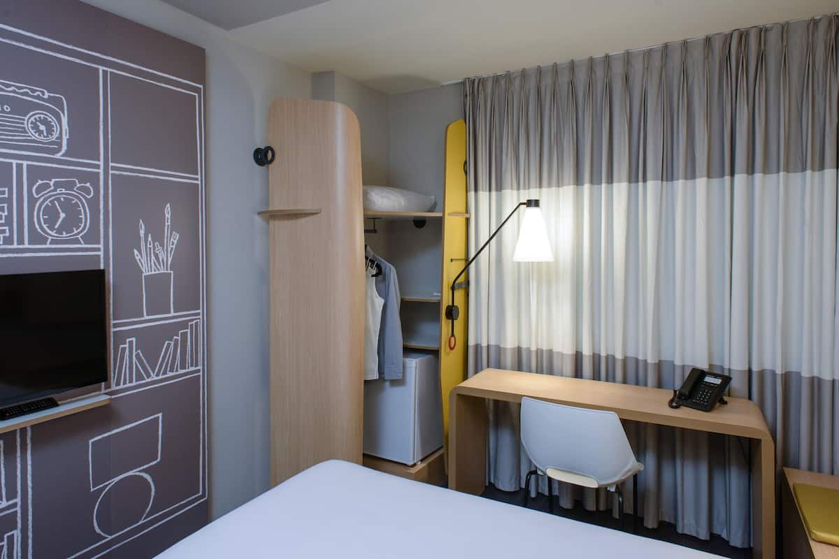 A hotel room with a bed, a desk with a chair, a TV, a wardrobe, and a window with curtains.
