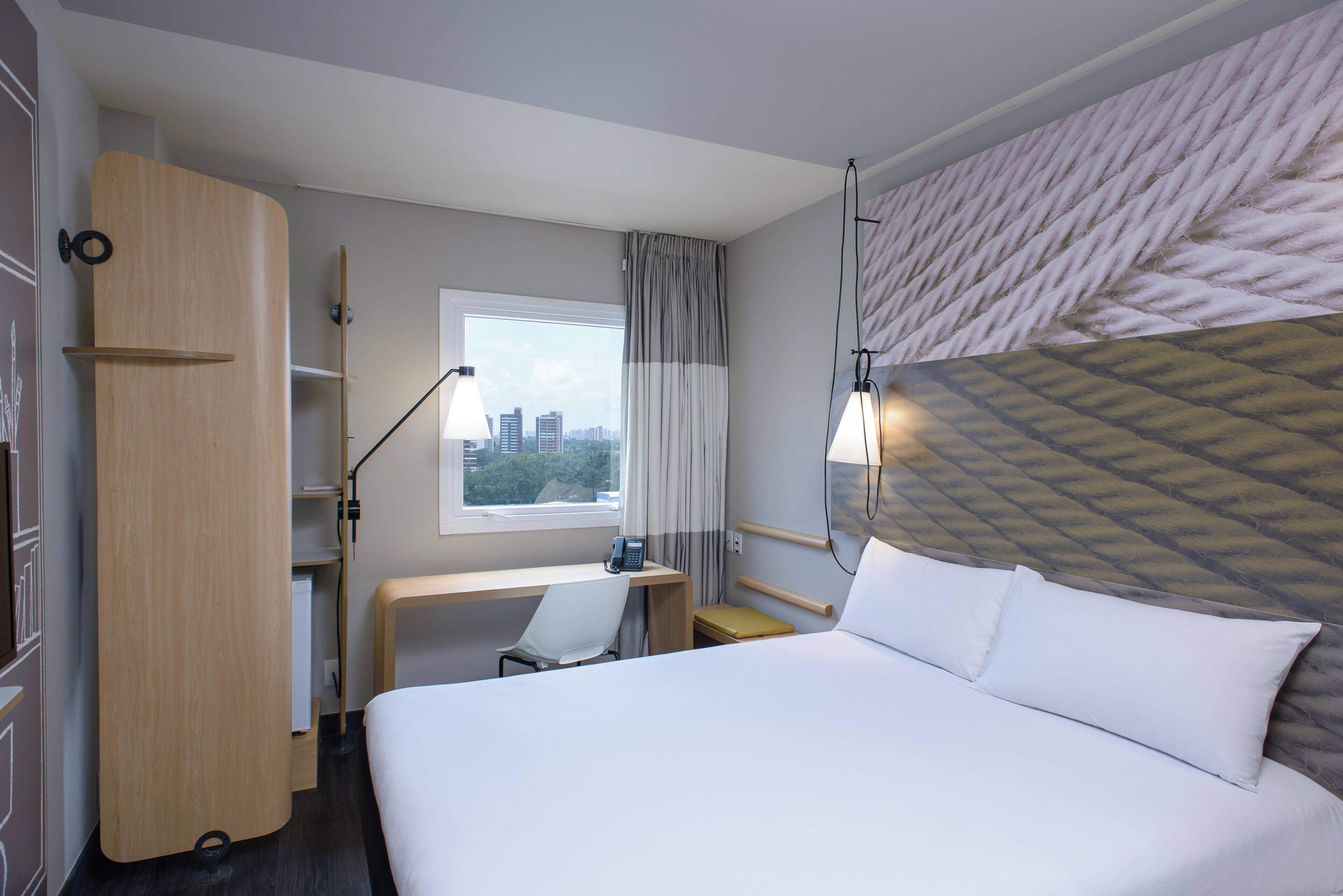 A hotel room with a bed, desk, chair, and a view of the cityscape through the window.