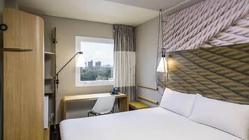 A hotel room with a bed, desk, chair, and a view of the cityscape through the window.