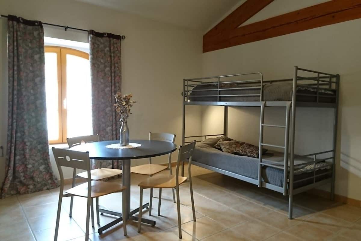 Een kamer met een stapelbed, een eettafel met stoelen, en een vaas met gedroogde bloemen.