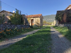 Un camí rural que condueix a una construcció de pedra amb teulada de teules, envoltada de vegetació i flors.