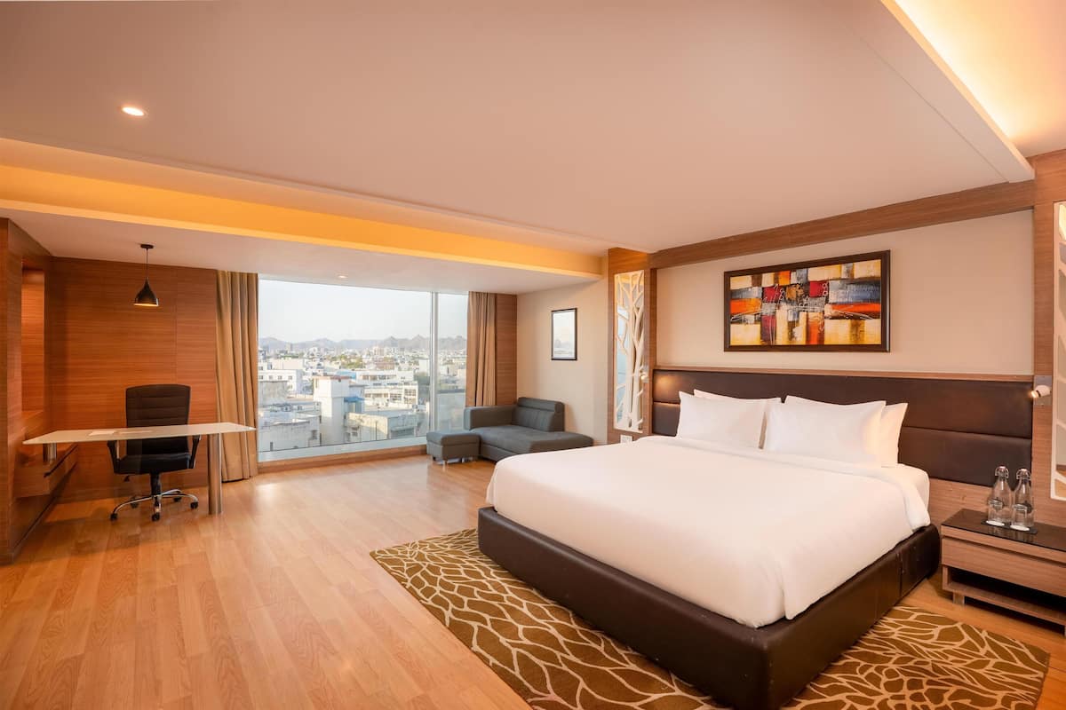 A hotel room with a large bed, a desk, a sofa, a city view, and a painting on the wall.