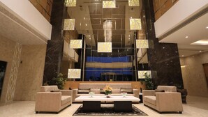 Eine großzügige Lobby mit modernem Design, inklusive einer zentralen Sitzecke mit Sofas und einem Couchtisch, einer großen Chandelier und einer Wand mit geometrischen Mustern.
