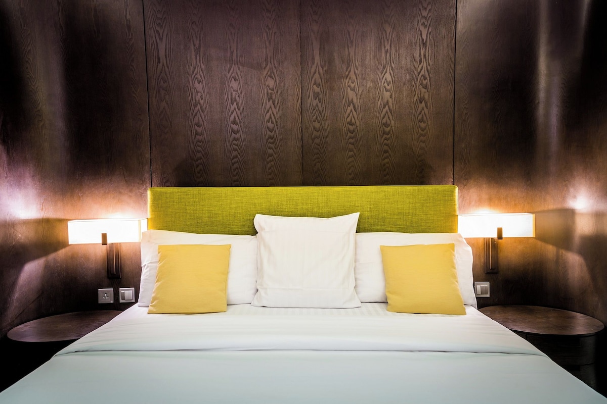 A hotel room with a wooden headboard, two bedside lamps, and a bed with white and yellow pillows.