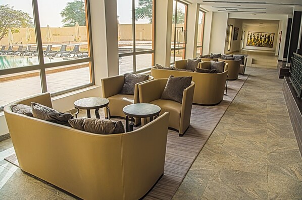 A modern lounge area with sofas and small round tables, large windows with a view of a swimming pool and outdoor seating.