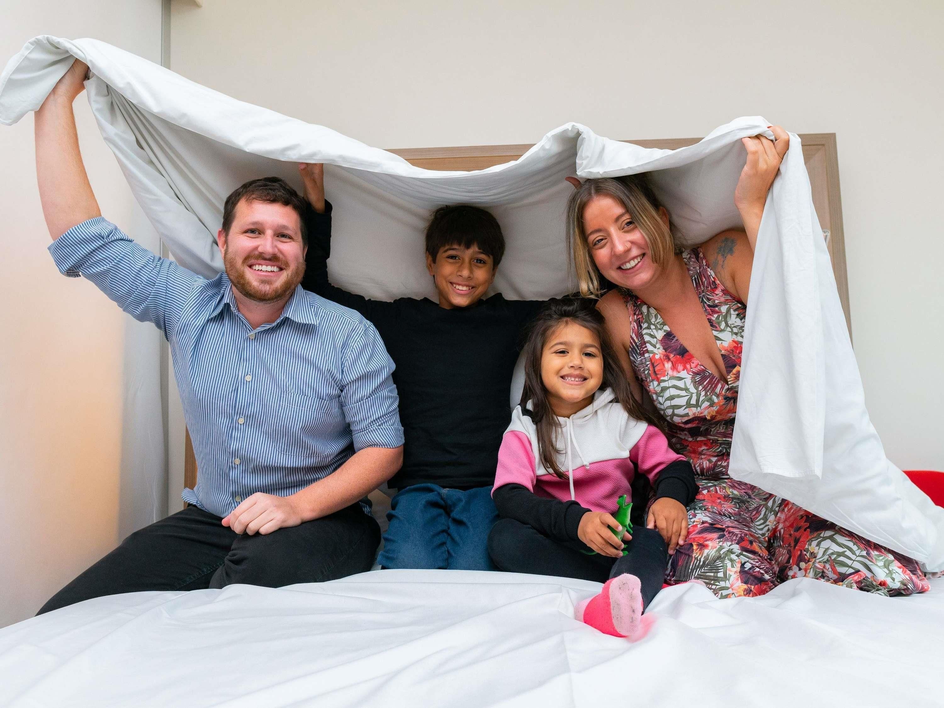 A family of four on a bed with a white sheet and a blanket.