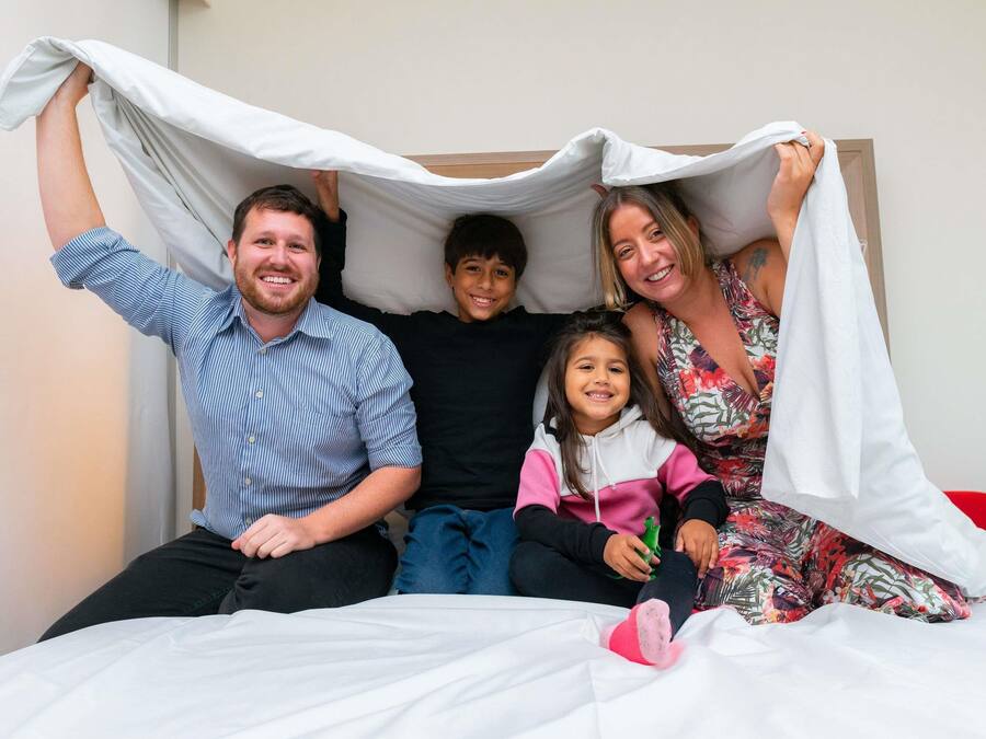 A family of four on a bed with a white sheet and a blanket.