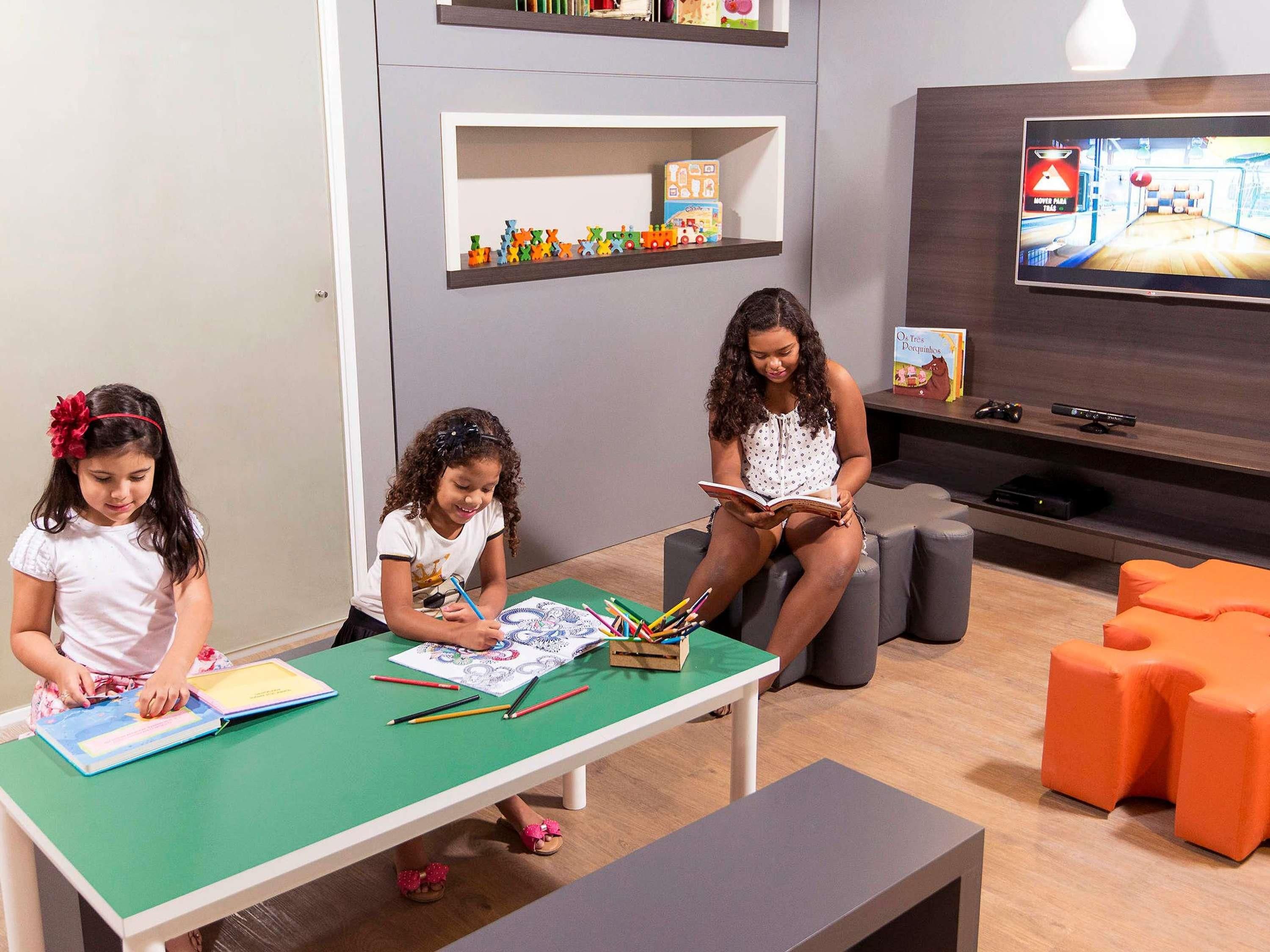 Una habitación con una mesa verde, dos niños dibujando, un televisor y una estantería llena de juguetes.