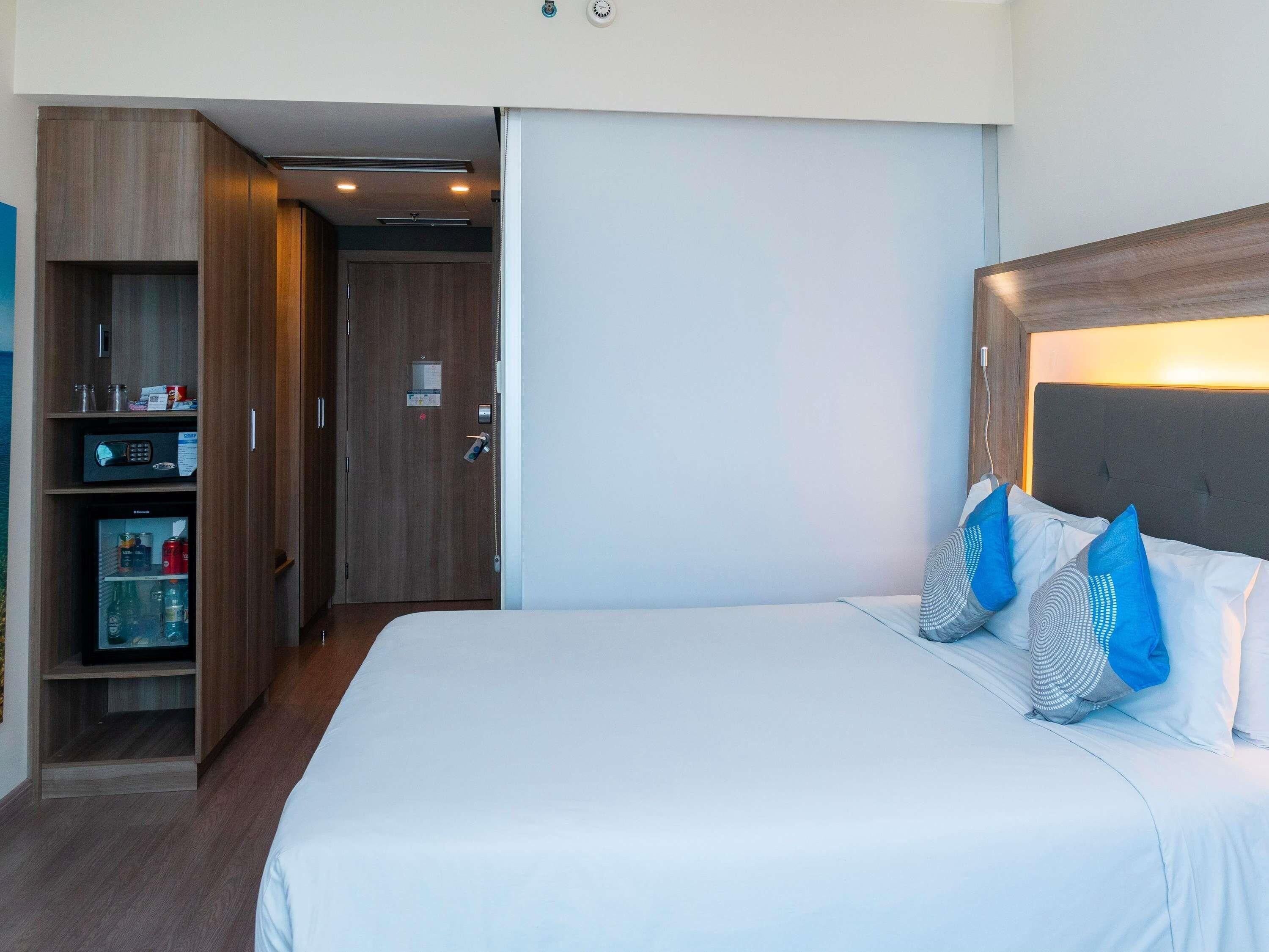 Una habitación de hotel con una cama grande, un armario, un minibar y una puerta que da a otra habitación.