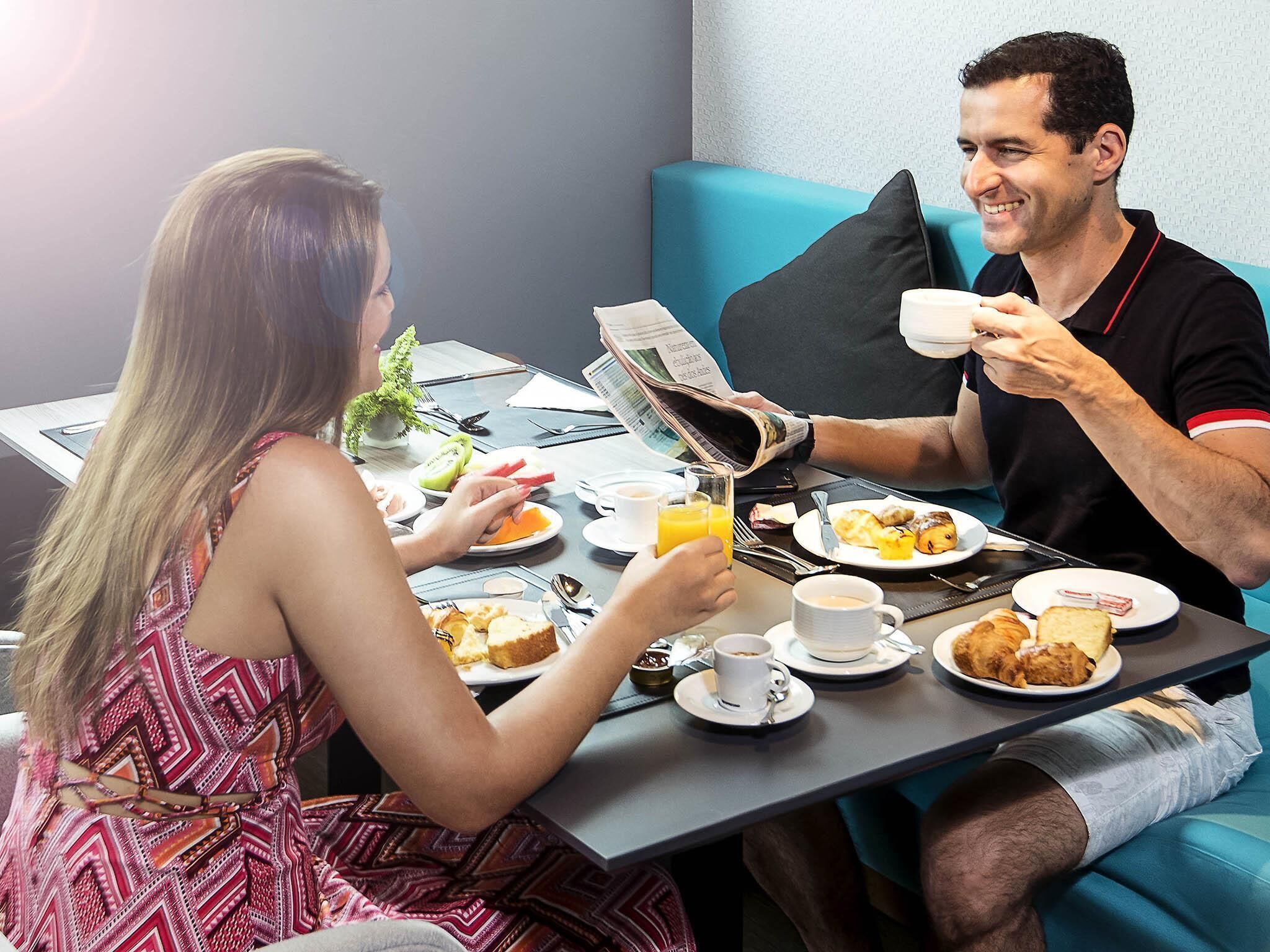 A breakfast setting with two people, a table with plates of food, cups, and a newspaper.