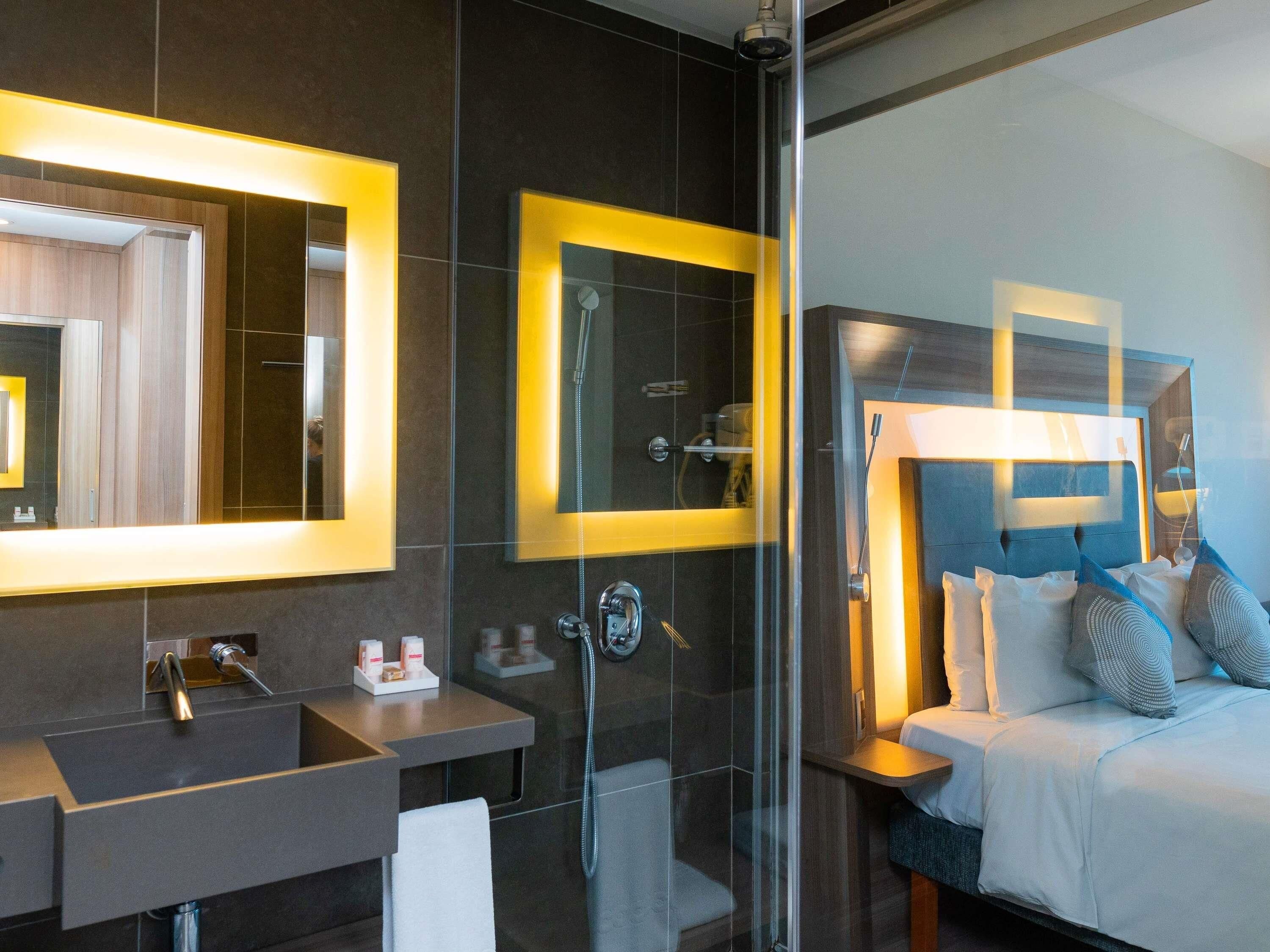 A modern bathroom with a glass shower enclosure, a vanity with a mirror, and a bed with pillows.