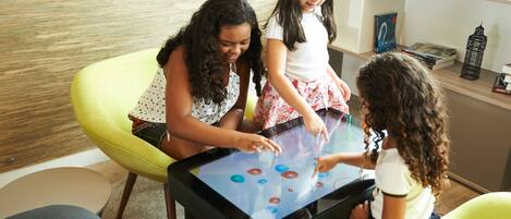 Three children playing with a digital interactive table in a modern room with a wooden floor and a bookshelf in the background.