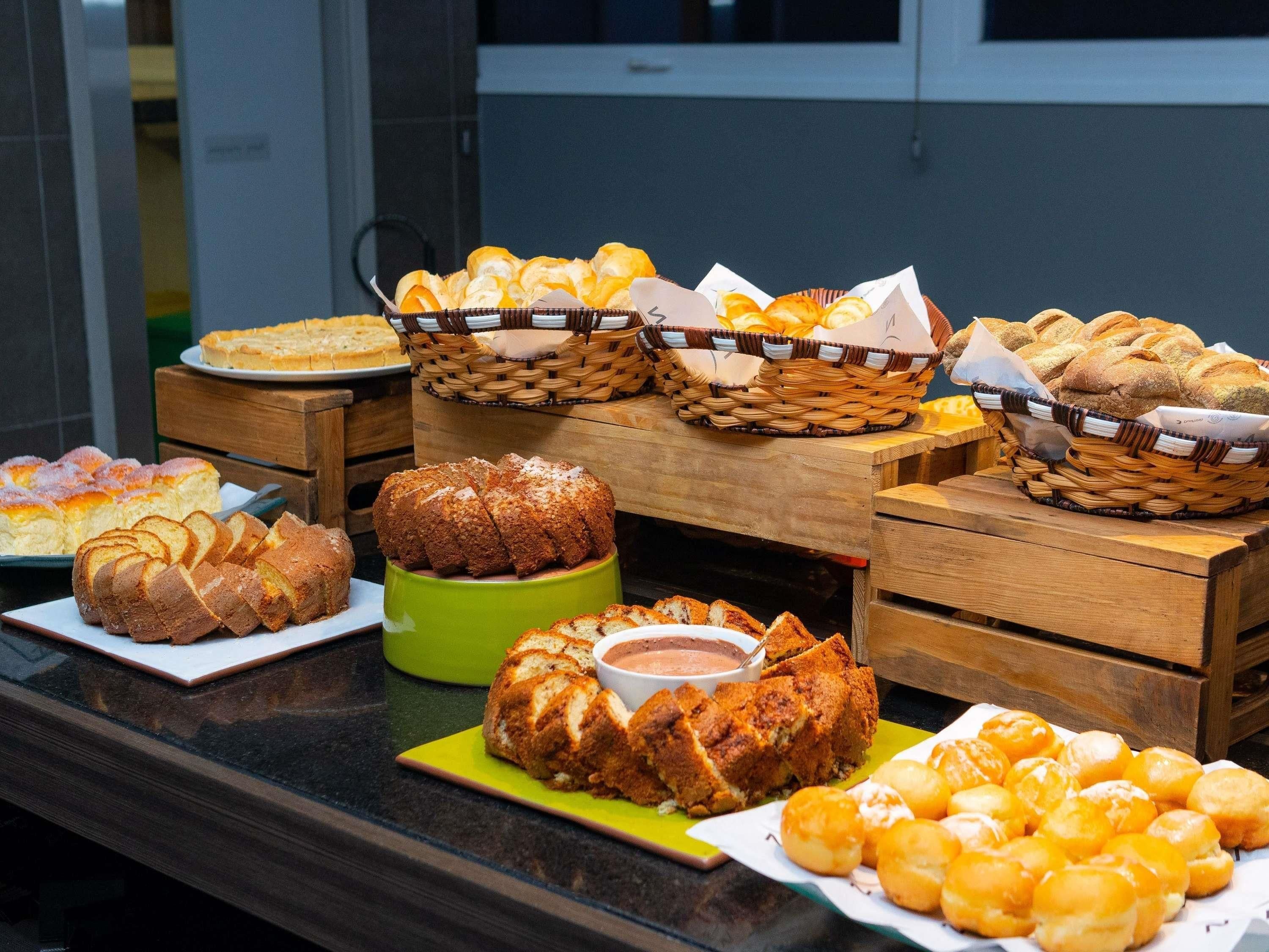 A buffet table with various breads and pastries, including sliced bread, rolls, and a loaf of bread in a green container.